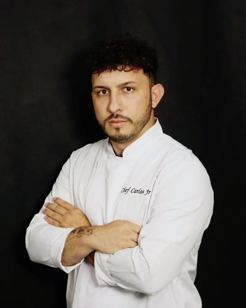 Chef Carlos Junior de dólmã branco, de braços cruzados, em fundo escuro